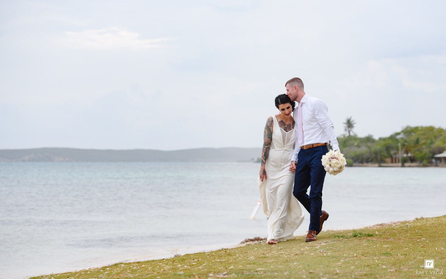 Bride and groom walking hand in hand along a tropical beach shoreline on their wedding day.