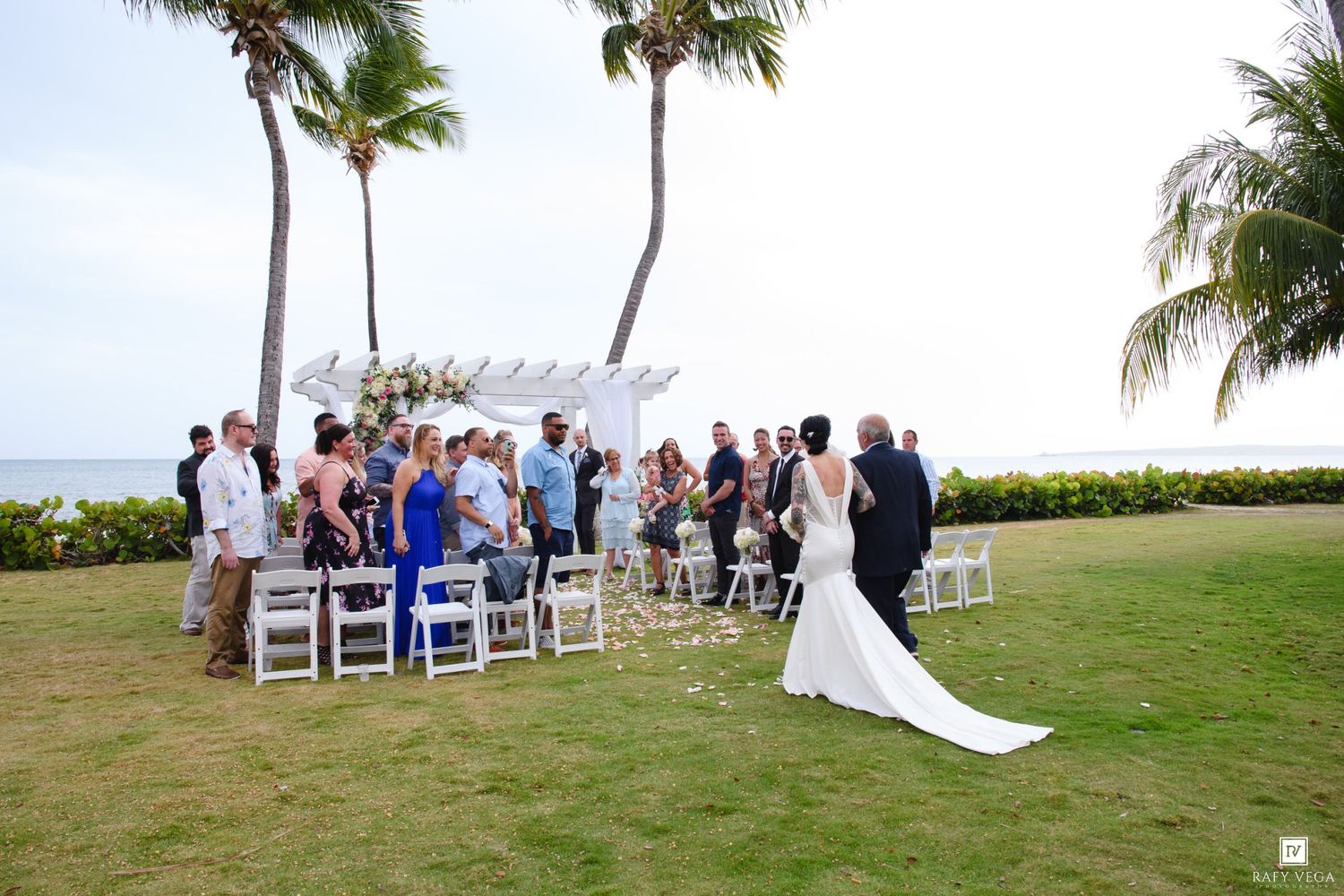 Outdoor beach wedding ceremony with bride in white gown, guests seated on white chairs under palm trees by the ocean.