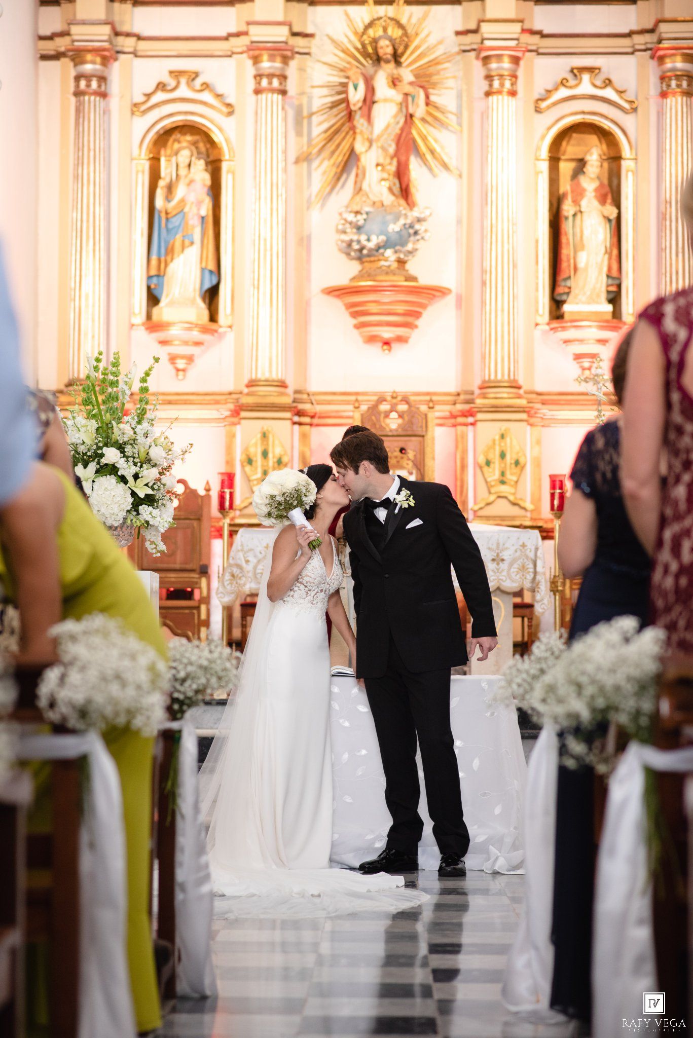 Bride and groom share first kiss at ornate church altar adorned with gold religious statues and white floral arrangements.