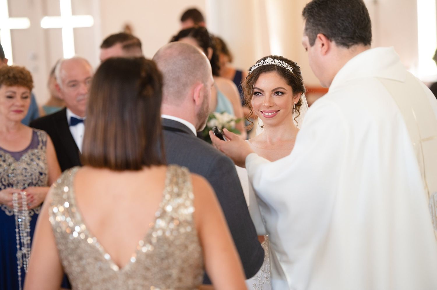 Bride smiling at groom during church wedding ceremony with officiant and guests in background.