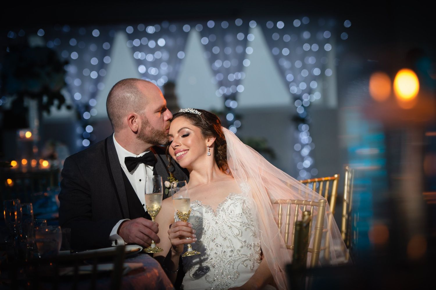 Bride and groom share a romantic moment with champagne glasses, posing before a sparkling fairy light backdrop at their wedding reception.