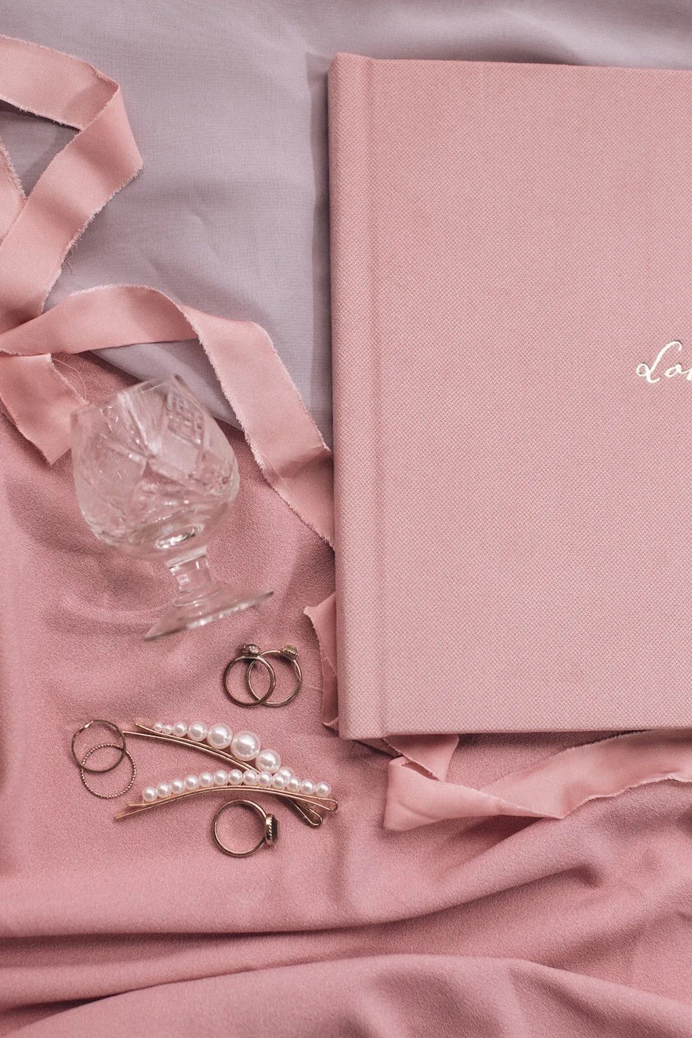 Pink hardcover love journal with pearl hair clips, rings, and crystal glass on pink silk fabric.