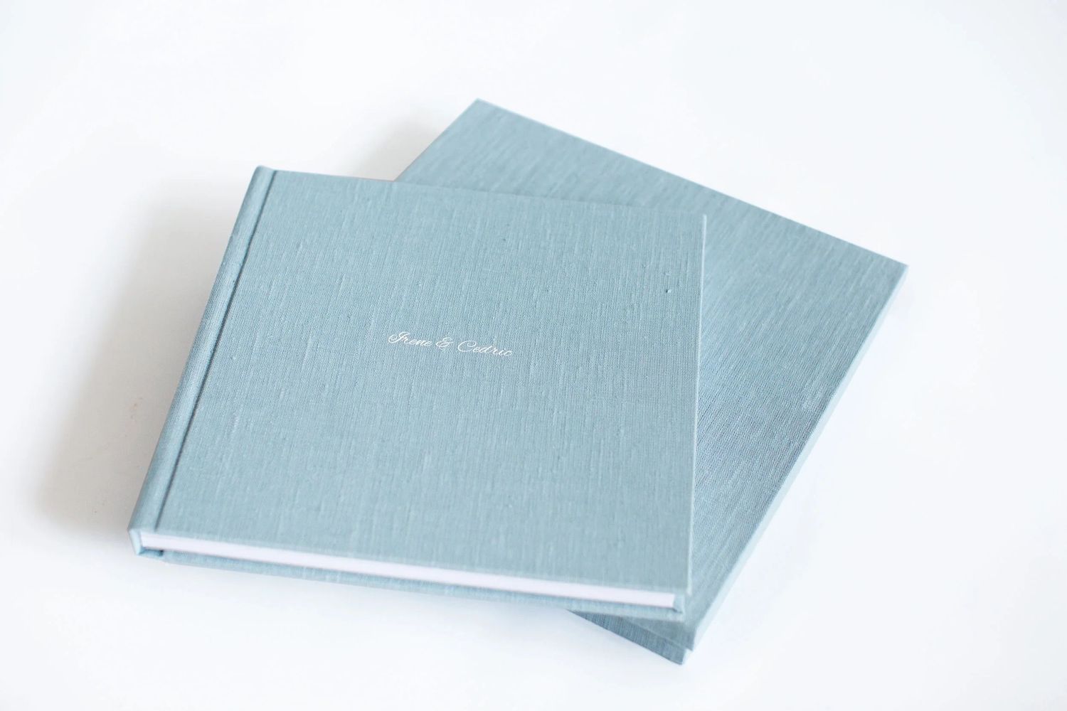Two elegant light blue linen hardcover wedding albums stacked on a white surface with gold foil text.
