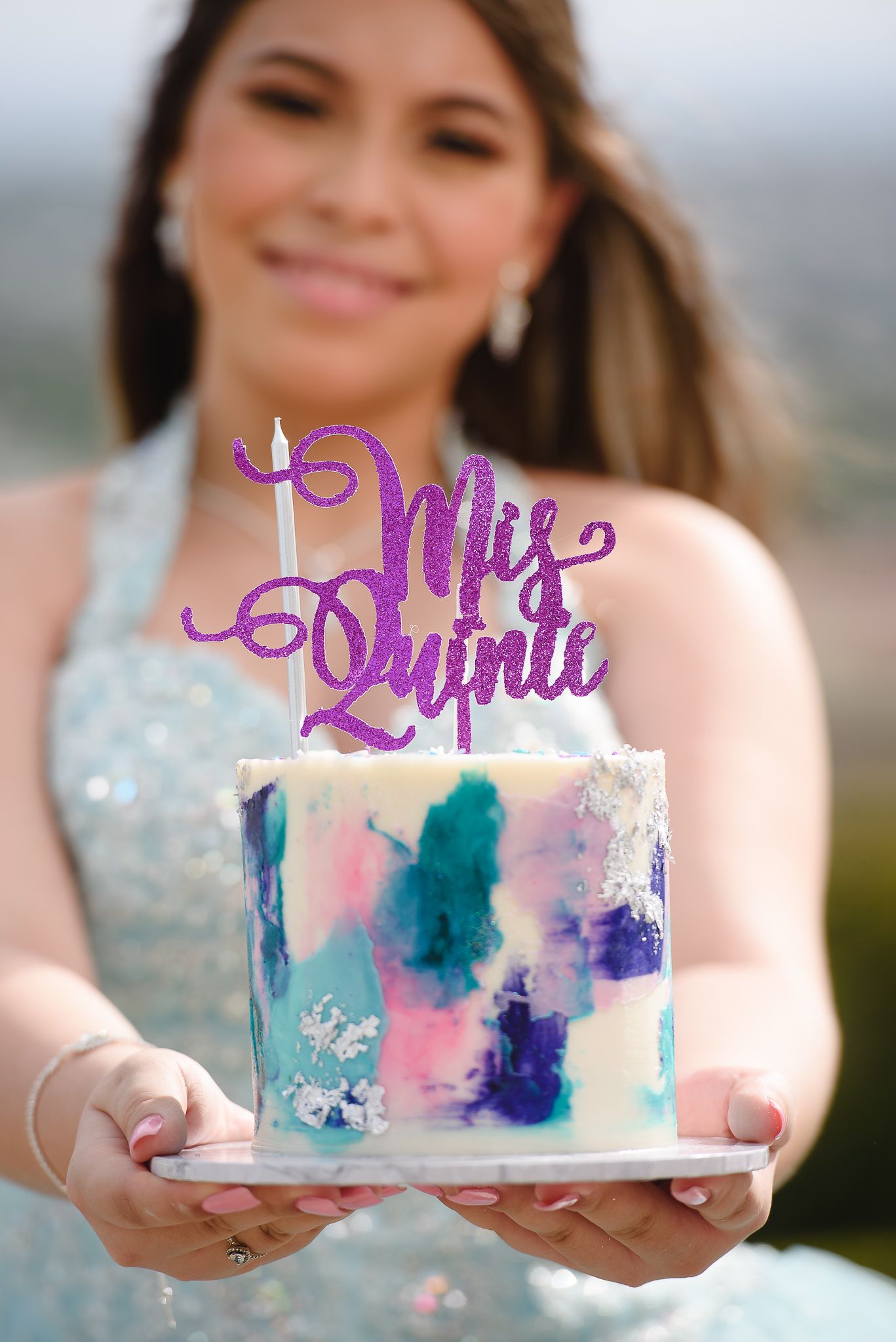 Young woman in blue dress holding a colorful watercolor Quinceañera cake with purple glitter topper outdoors.