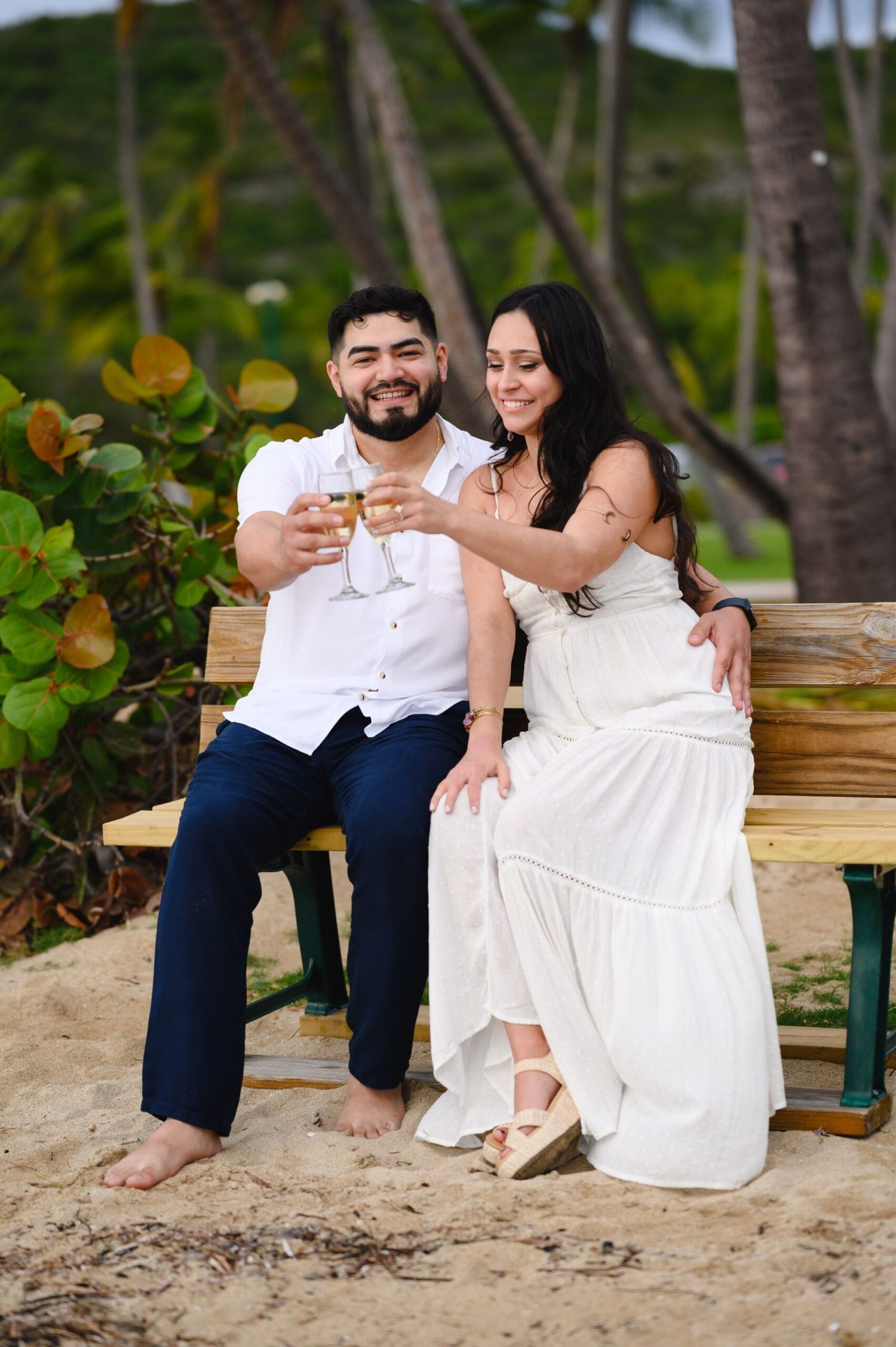 Couple toasting champagne on a beach bench, man in white shirt and navy pants, woman in white dress, palm trees behind.