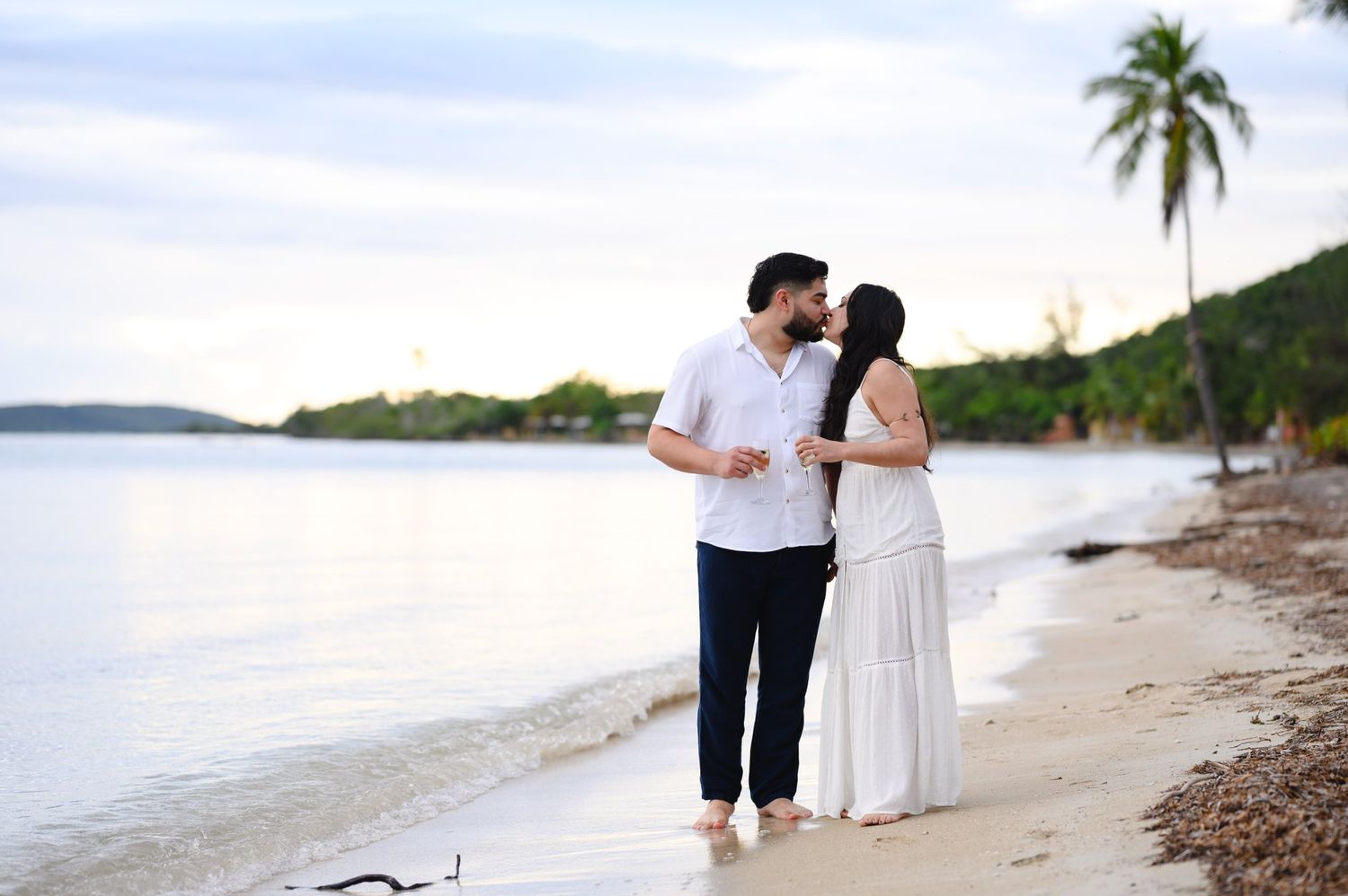 Couple kissing on a tropical beach at sunset, woman in white dress, man in navy pants, palm trees and calm water.