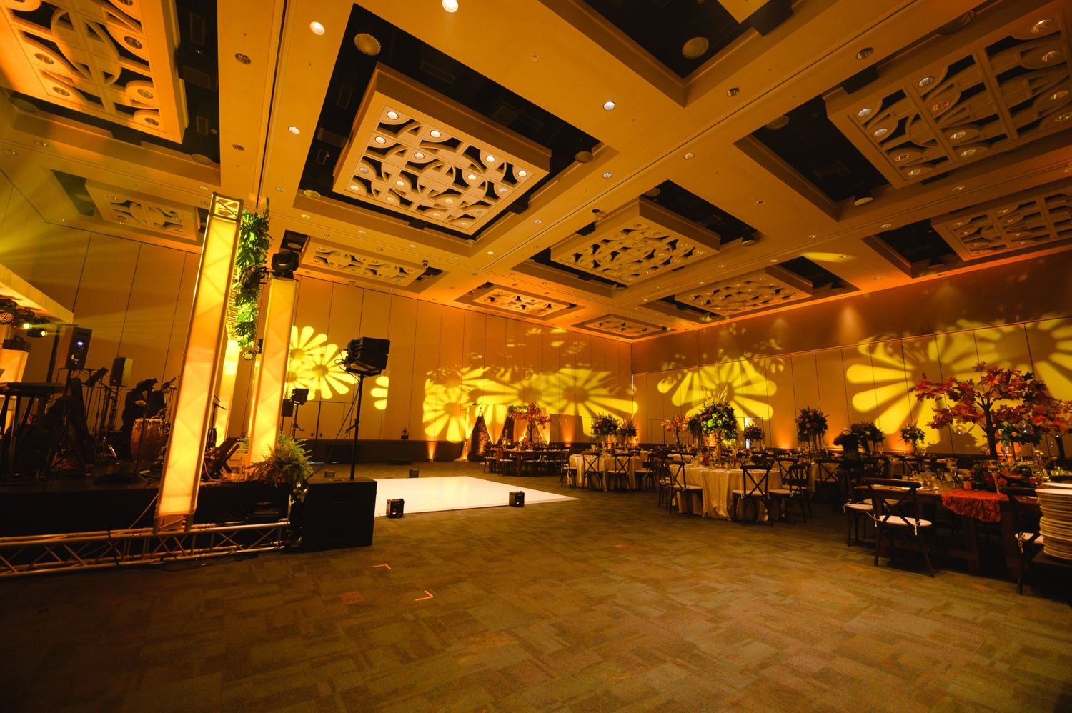 Elegant ballroom with golden uplighting, decorative ceilings, palm projections, and round dining tables for an upscale event.
