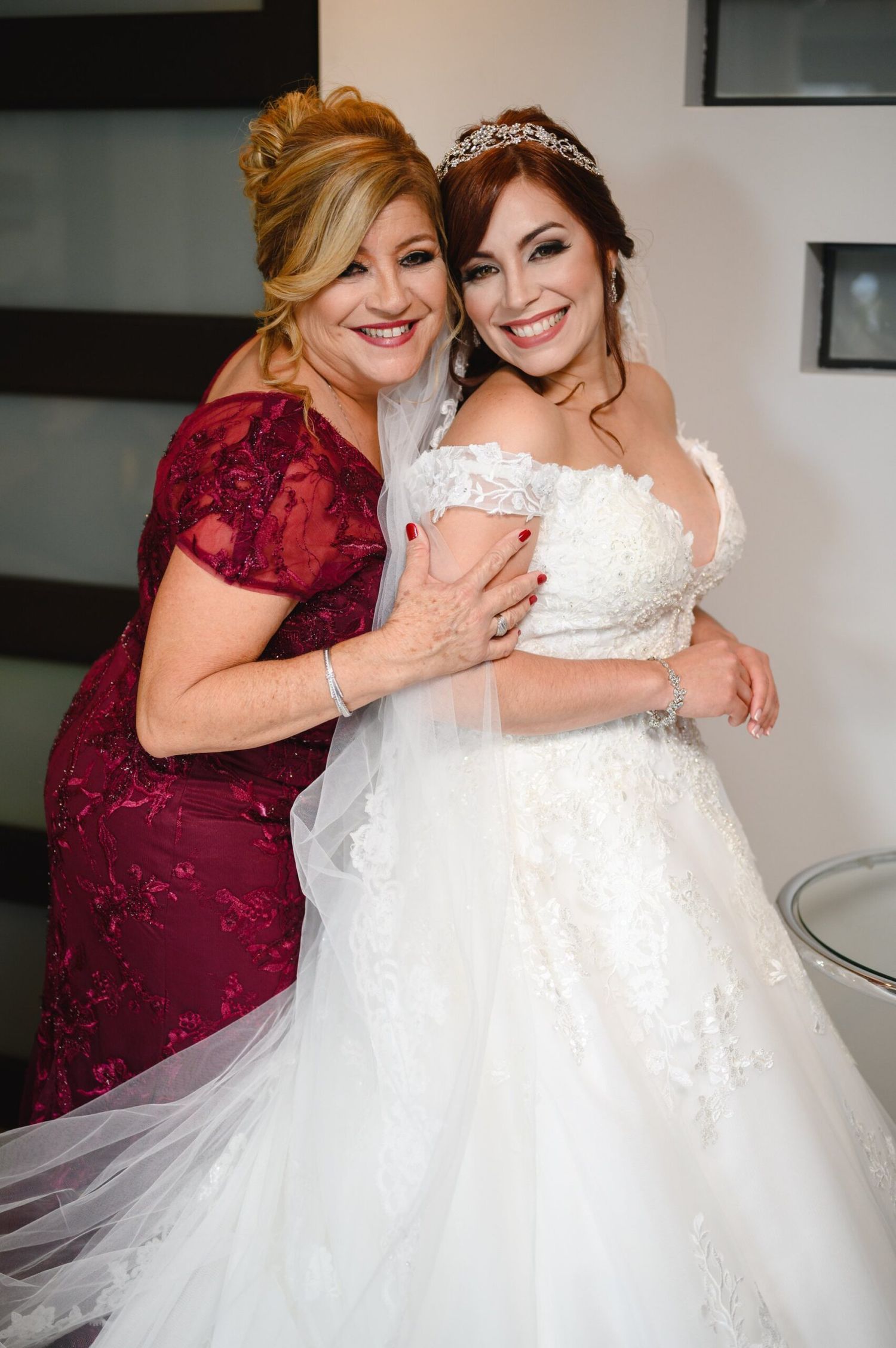Bride in white lace gown with tiara embraces woman in burgundy dress, both smiling joyfully on wedding day.