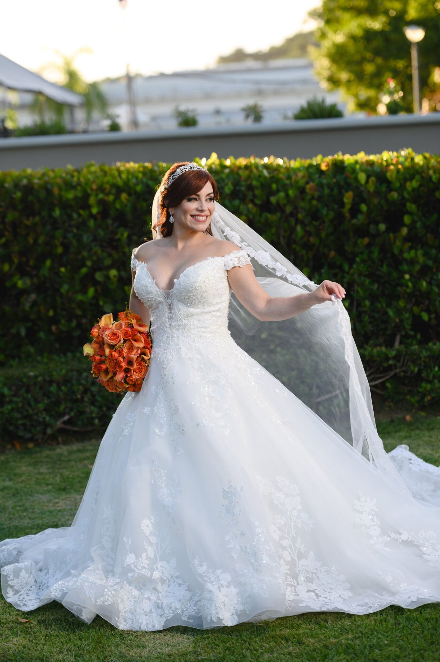 Bride in off-shoulder lace ball gown wedding dress holding orange bouquet and veil outdoors.