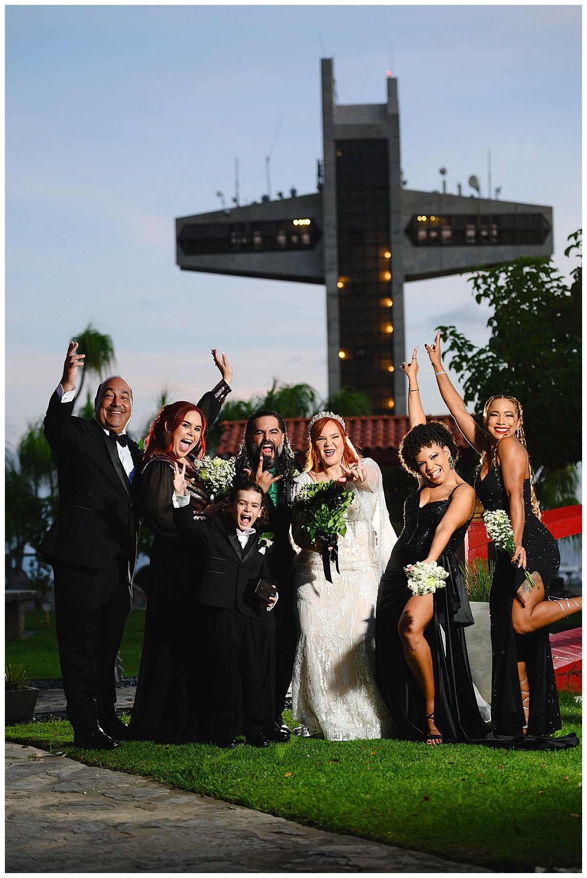 Wedding party celebrating outdoors with bouquets raised, bride in white gown, guests in black formal wear, large cross monument behind.