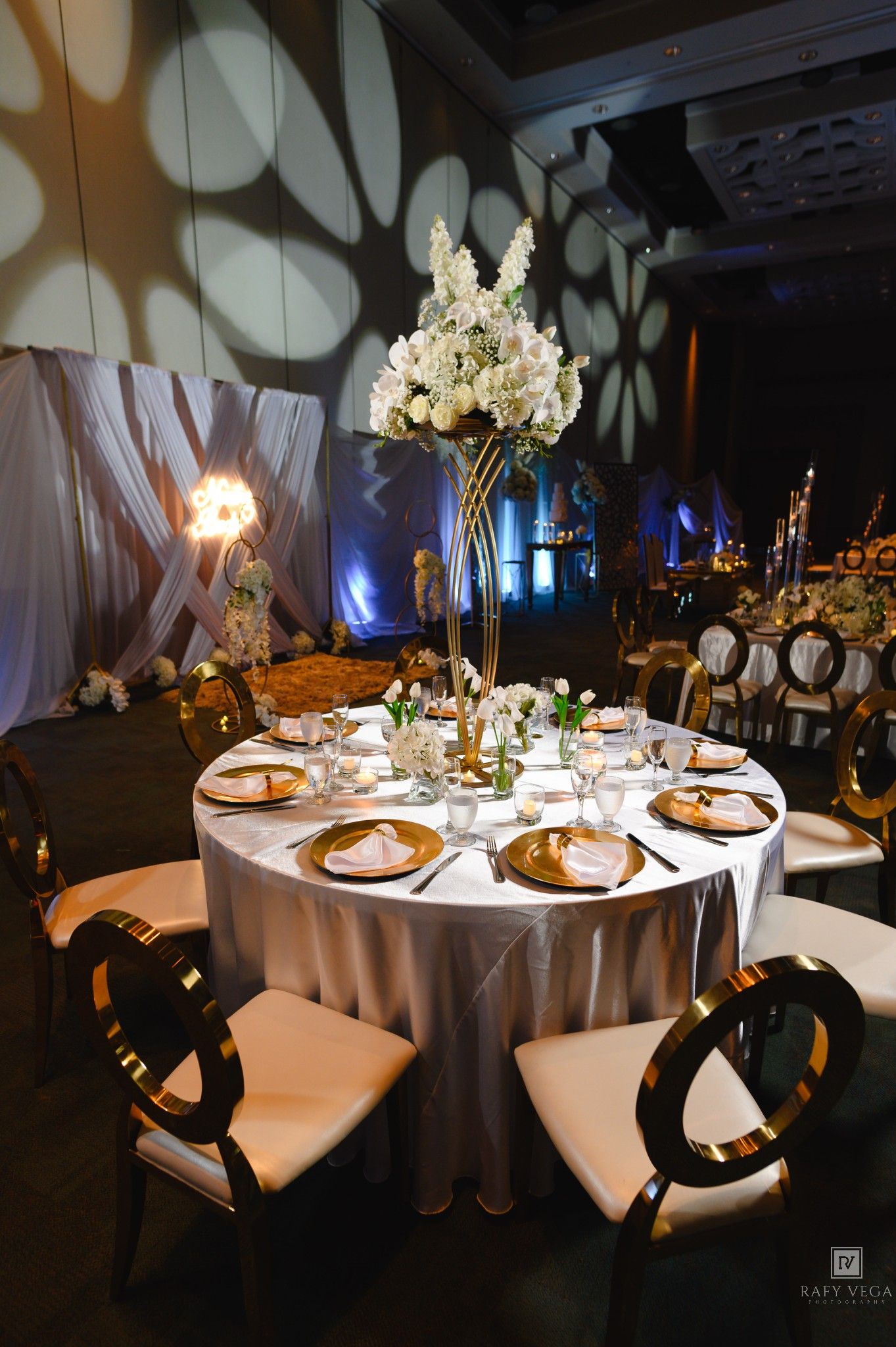 Luxurious wedding reception table with gold chargers, tall floral centerpiece, and dramatic lighting.