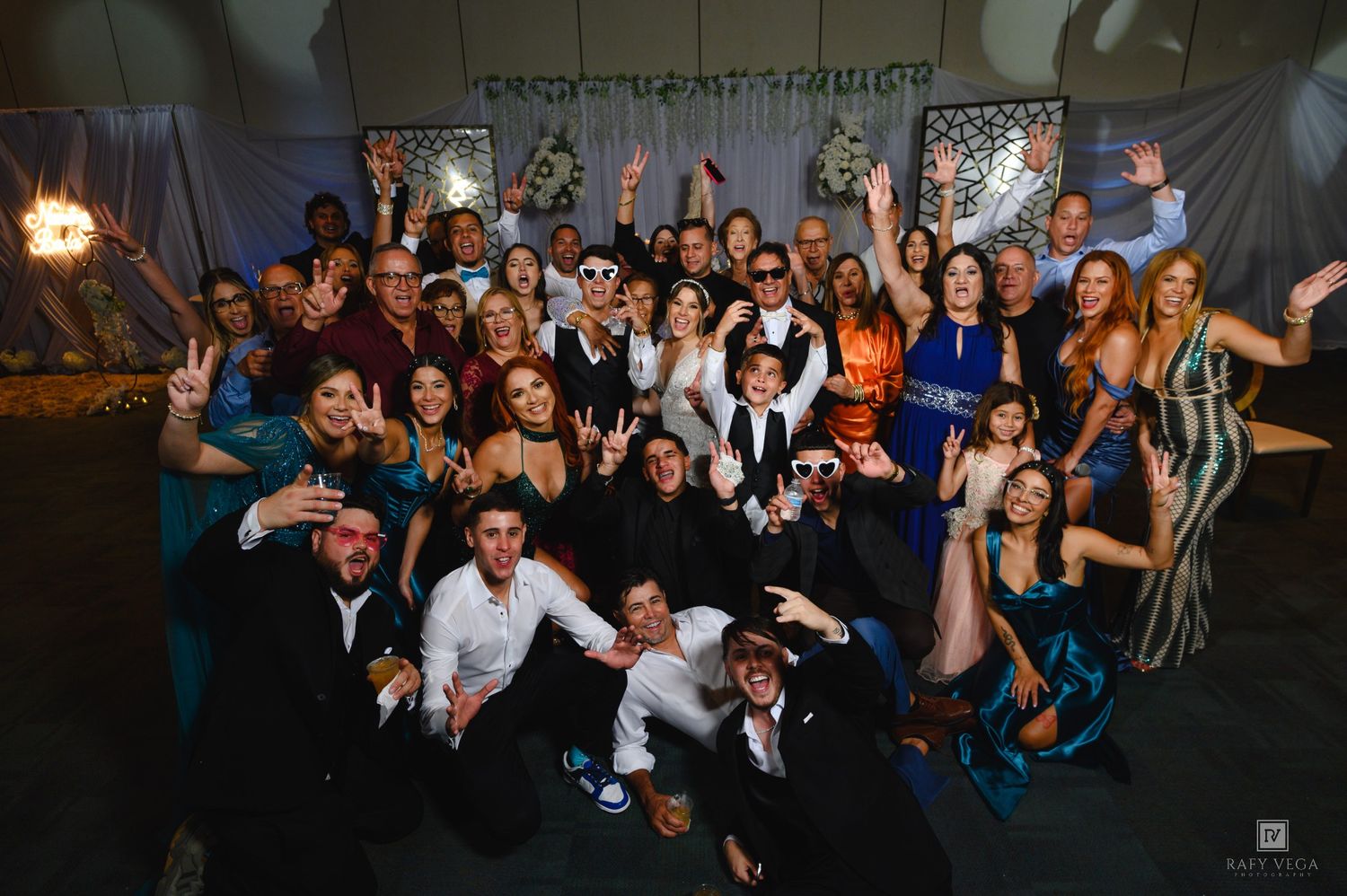 Large group of elegantly dressed people posing together at a formal gala event with decorative backdrop.