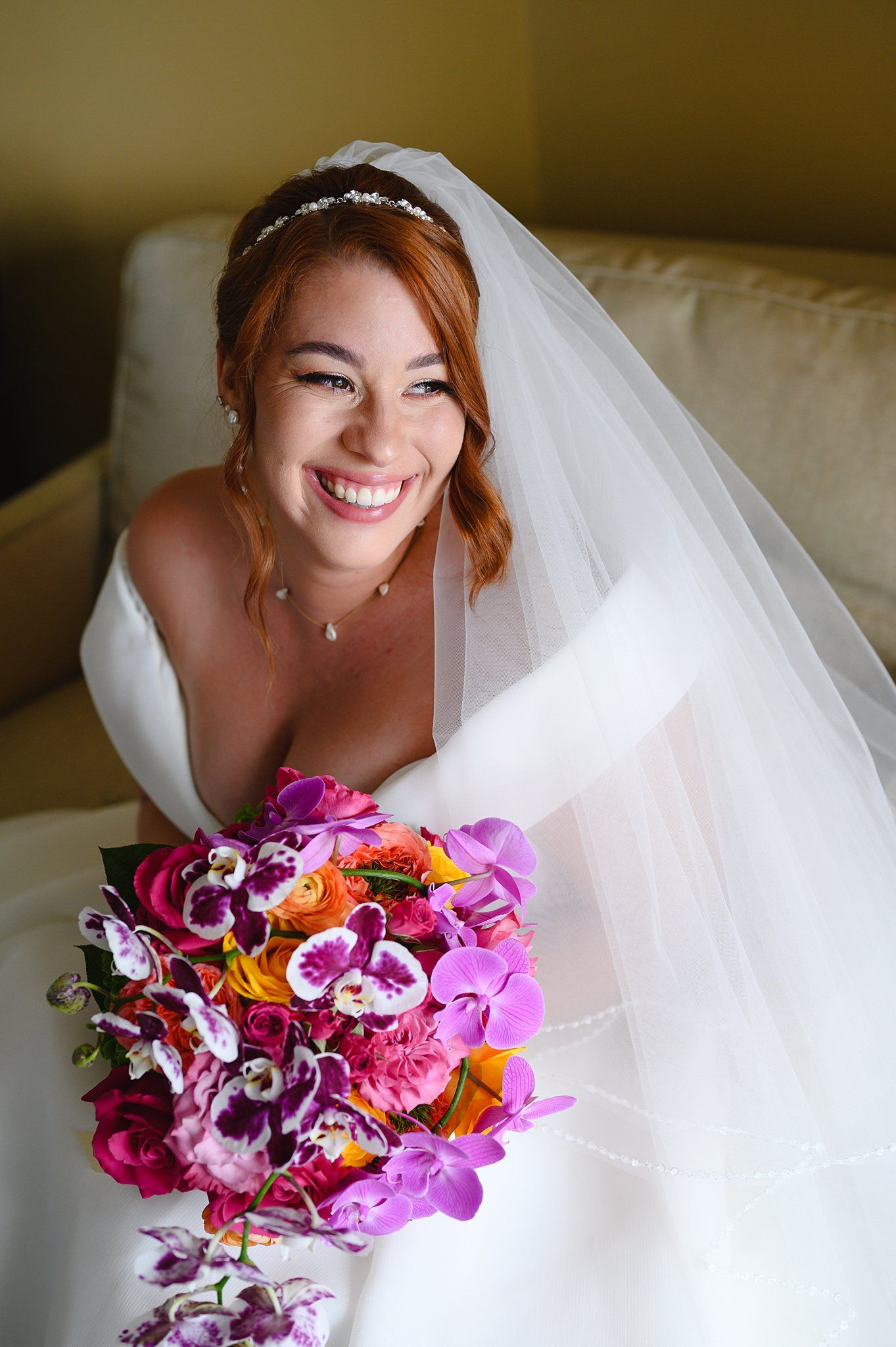 Smiling bride in white gown and veil holds vibrant purple and pink orchid bouquet while seated on a couch.