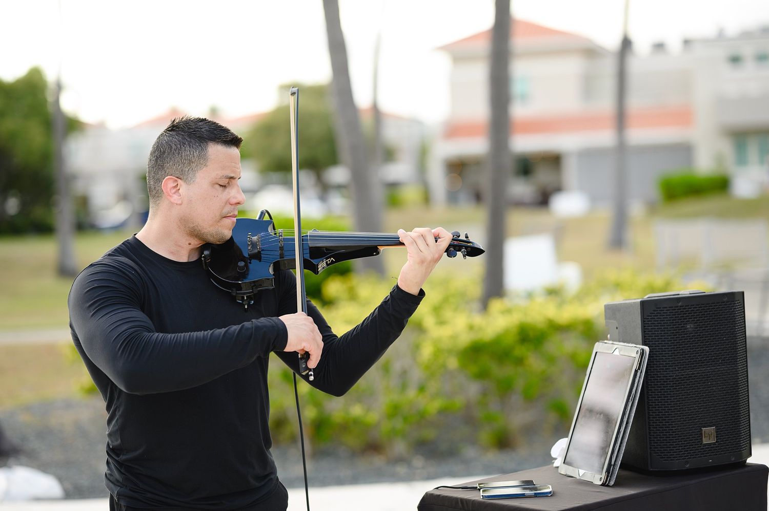Man in black playing a blue electric violin outdoors at an event with a speaker and laptop nearby.