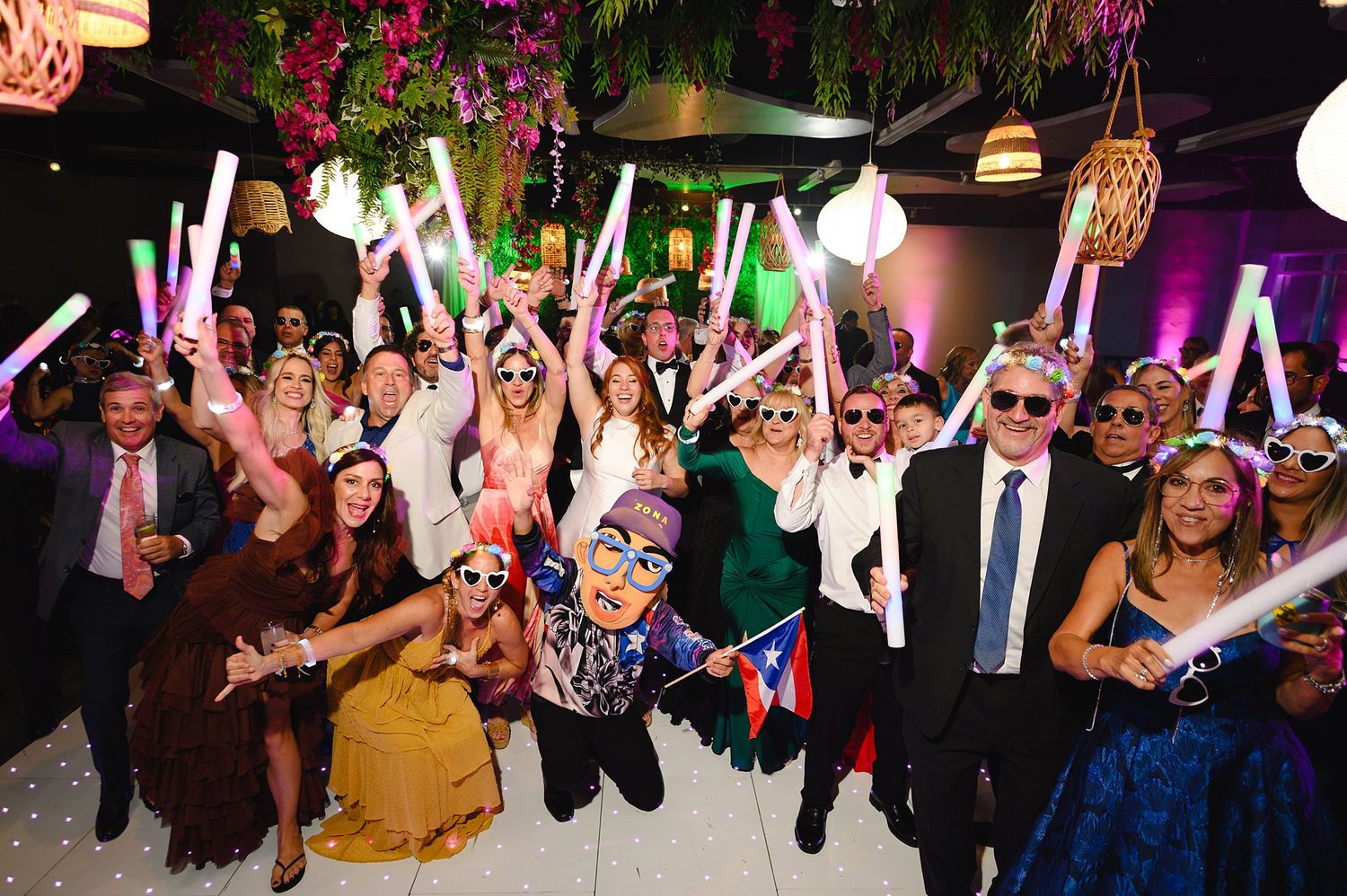Wedding guests celebrating with glowing light sticks and fun glasses at a lively indoor party reception.