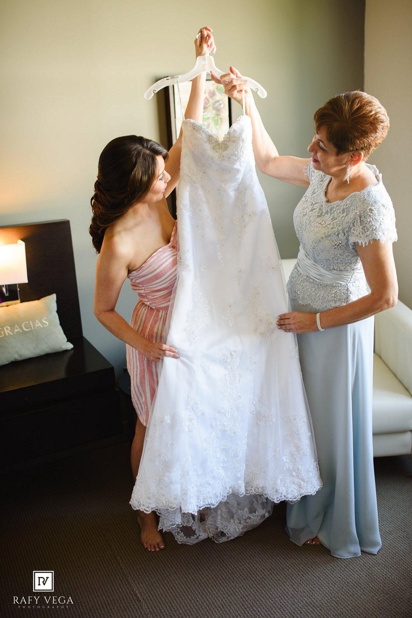 Two women holding up a white lace wedding dress on a hanger in a hotel room before the ceremony.