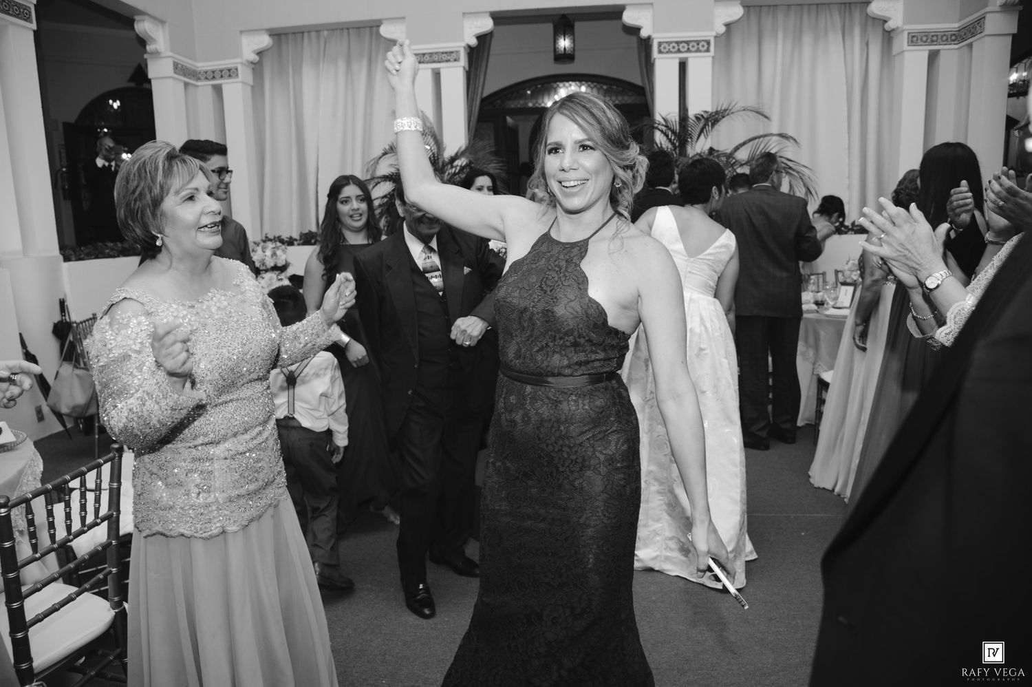 Black and white photo of elegantly dressed women dancing joyfully at a formal wedding reception event.