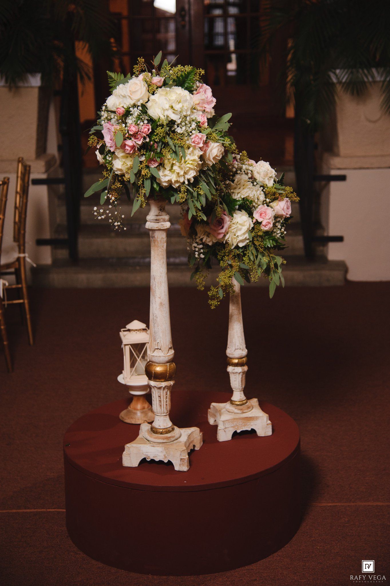 Elegant wedding floral arrangement with pink and white roses on ornate gold candlestick stands atop a burgundy platform.