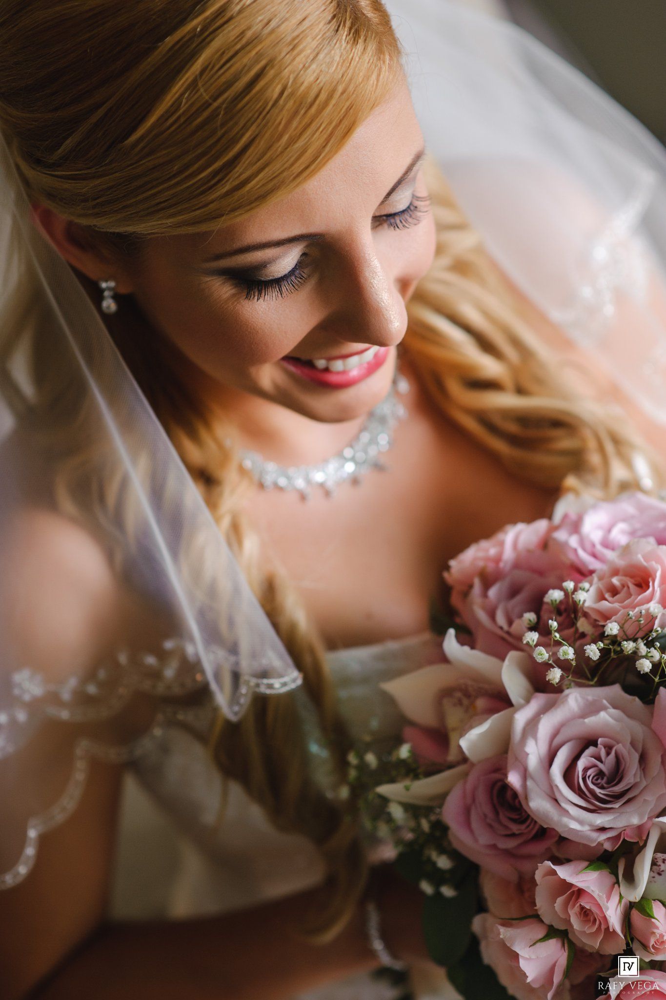 Beautiful blonde bride smiling down at her pink rose bouquet wearing a veil and crystal necklace on her wedding day.
