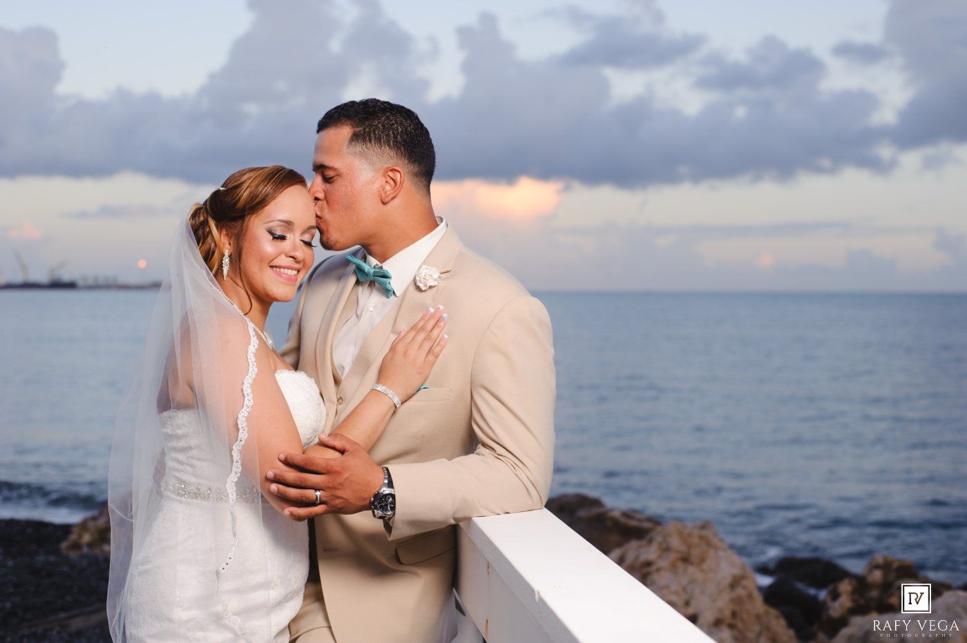 Groom in beige suit kisses bride in white gown and veil on oceanfront balcony at sunset wedding.