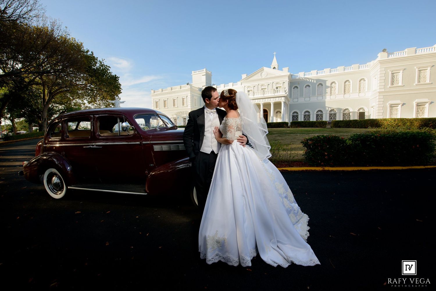 Bride and groom kissing beside a vintage dark red car with a white colonial building in the background.