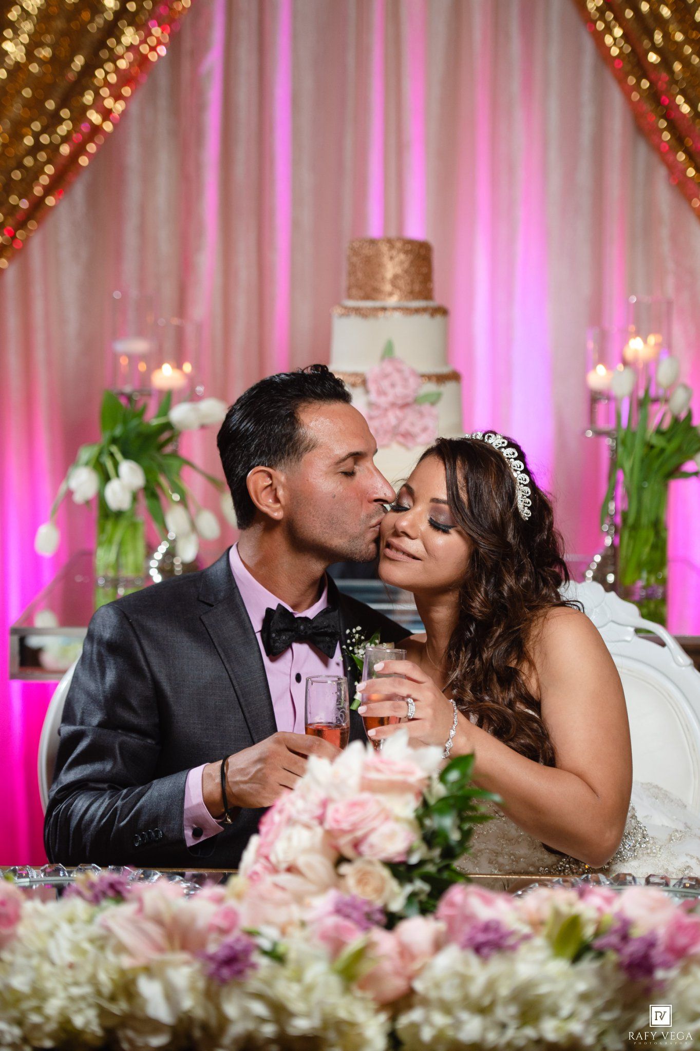 Bride and groom share a kiss at their wedding reception, surrounded by pink florals and a white tiered cake.