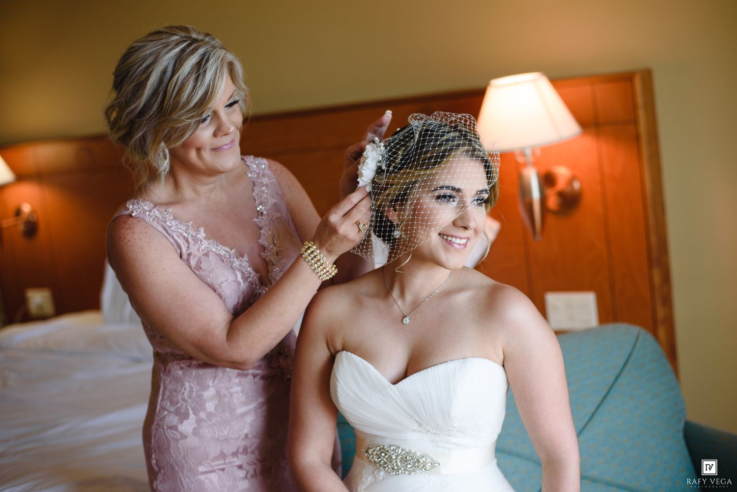 Bride in white strapless gown gets birdcage veil adjusted by woman in pink lace dress in hotel room.