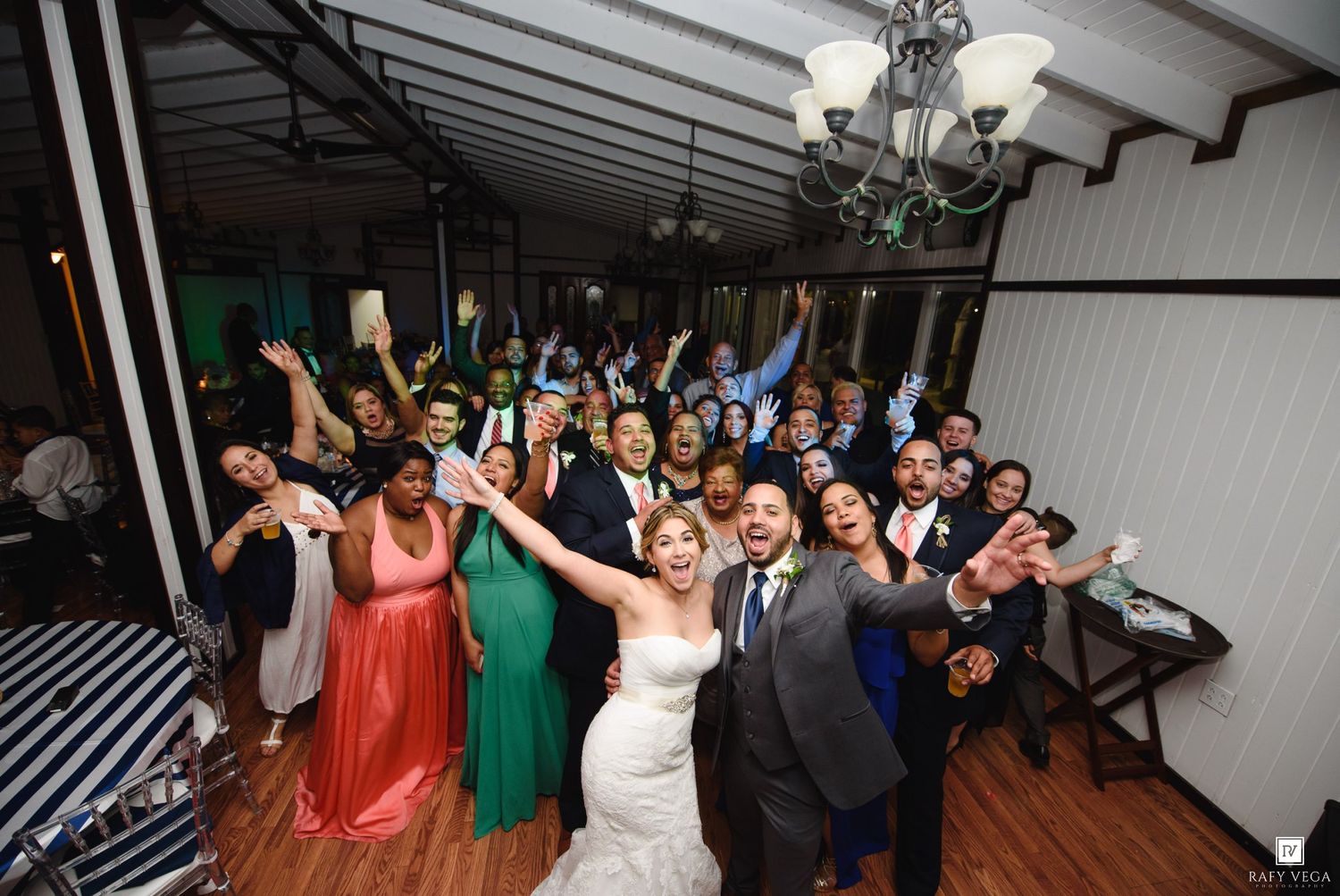 Joyful wedding reception group photo with bride in white gown and guests celebrating together indoors.