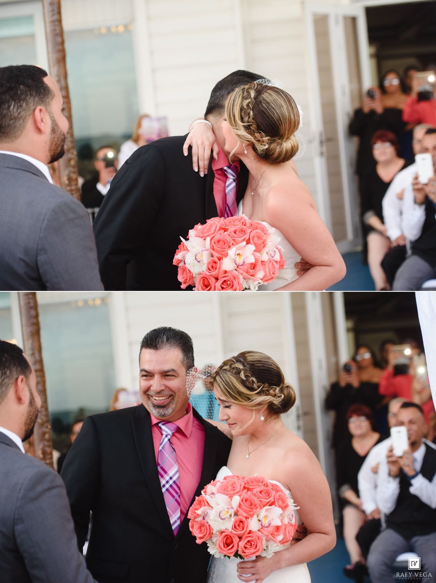 Bride holding coral rose bouquet shares a kiss with groom in black suit at outdoor wedding ceremony.