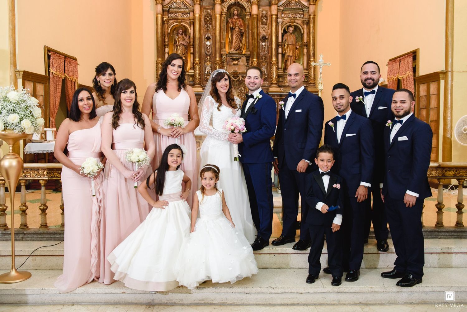Wedding party poses together in church, bridesmaids in blush gowns with groomsmen in navy suits.