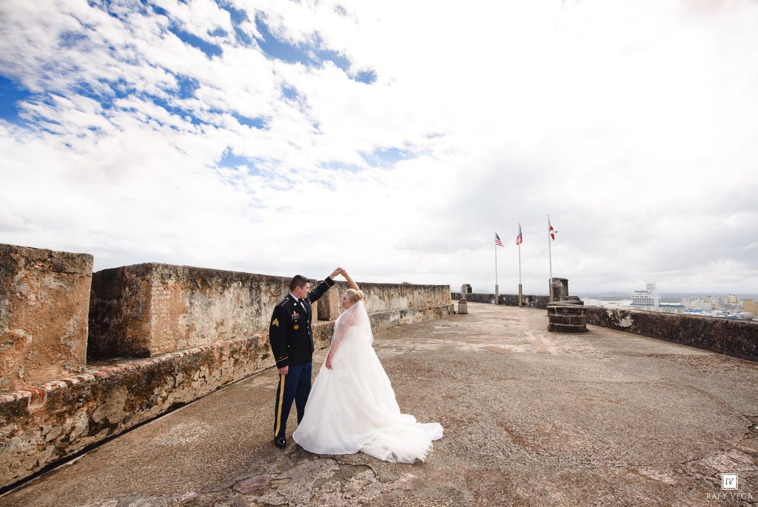Bride and groom share a romantic dance on a historic stone fortress terrace under a blue sky with flags in the background.