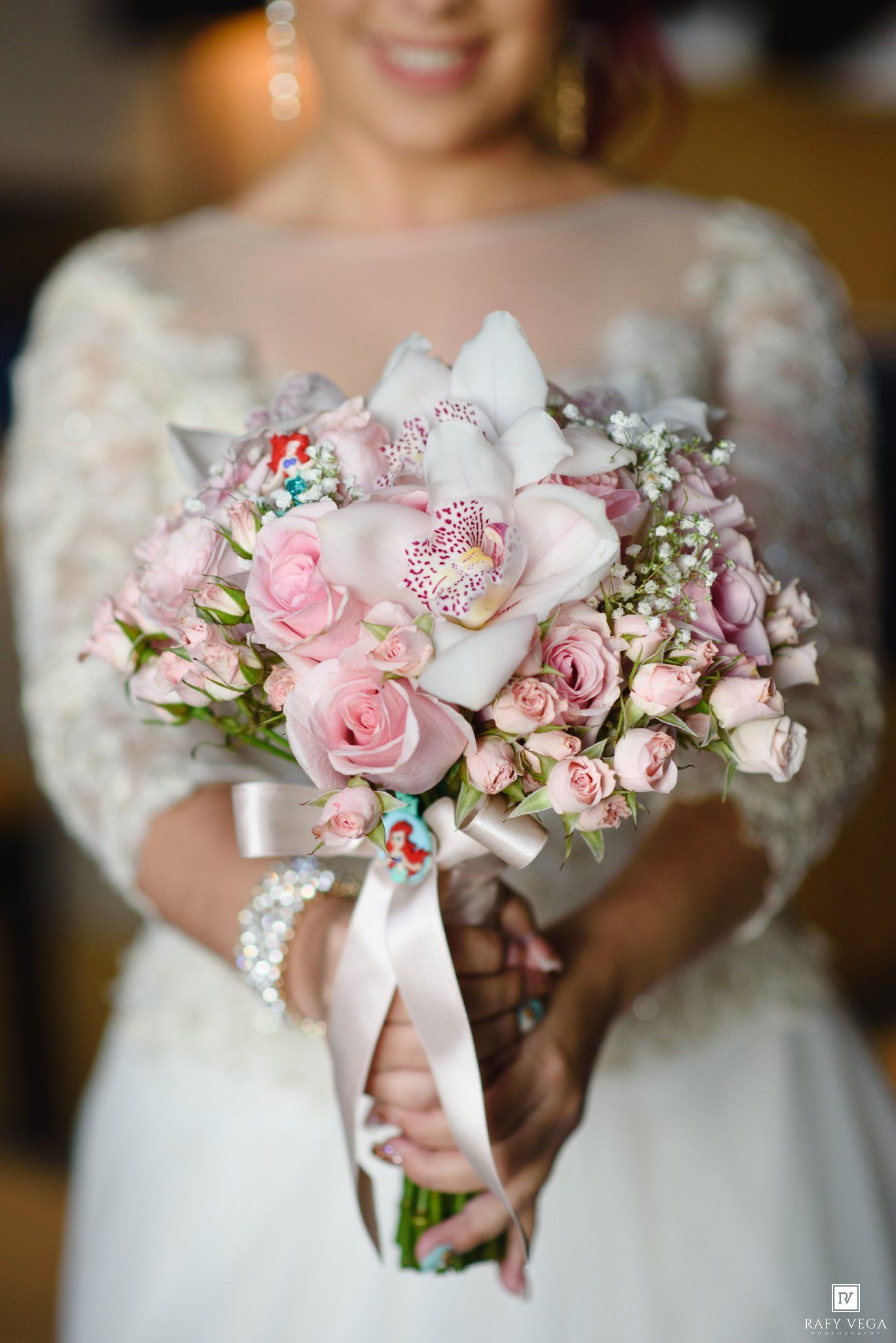 Bride holding a romantic pink bridal bouquet with roses and orchids, tied with a satin ribbon, wearing a white lace gown.