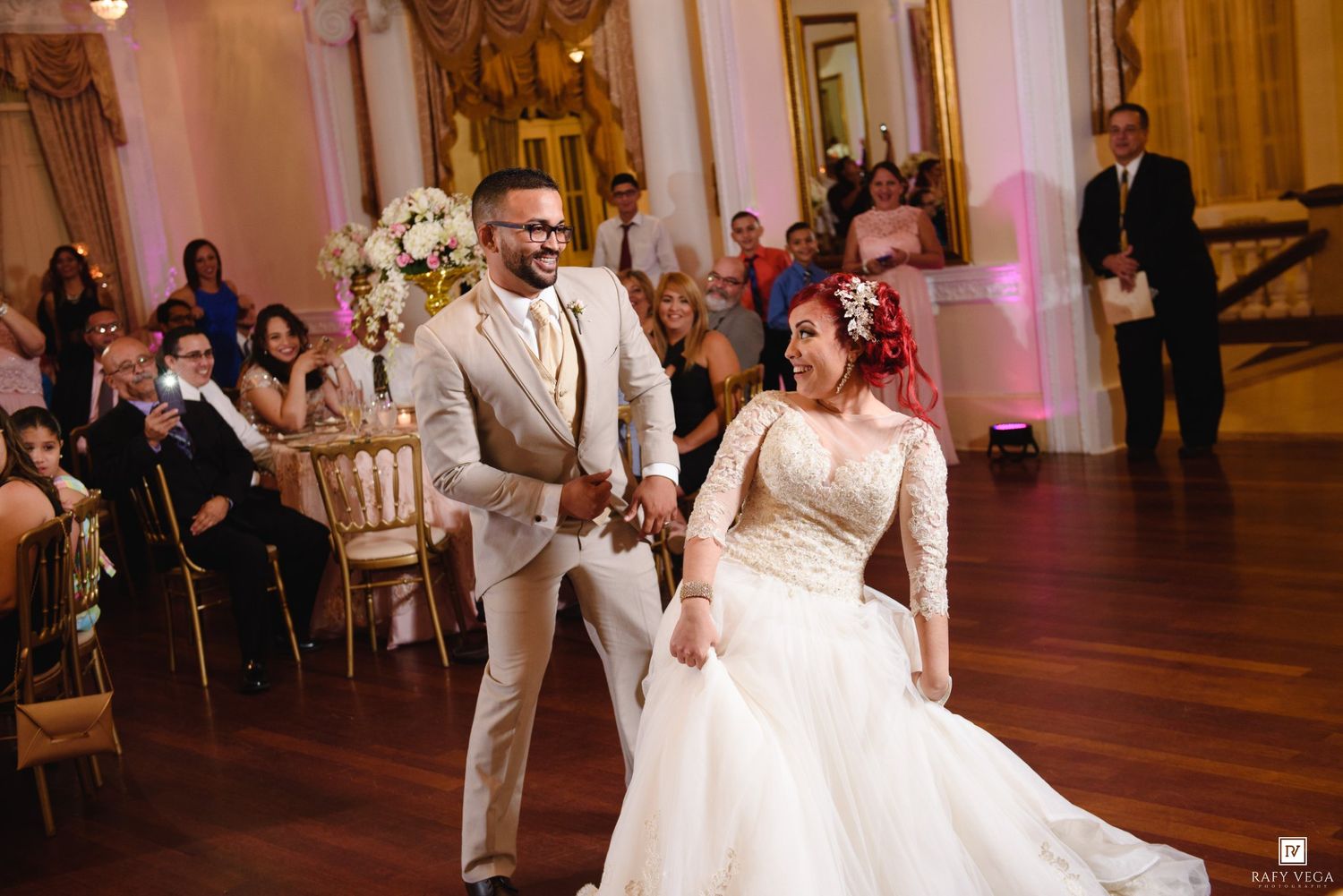 Bride in white lace gown and groom in tan suit share first dance at elegant ballroom wedding reception.