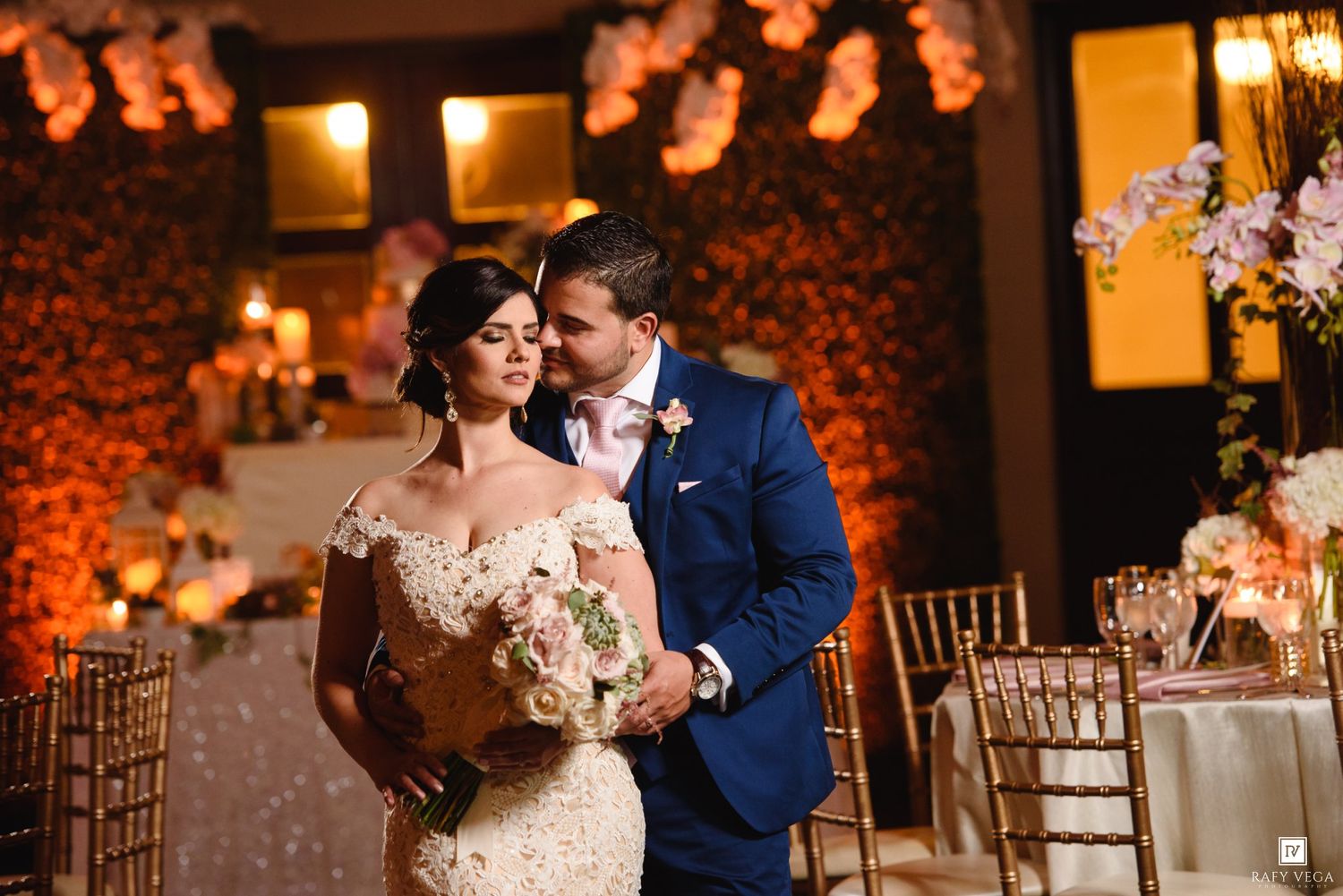 Bride and groom share romantic moment at elegant wedding reception with warm floral backdrop and gold chiavari chairs.