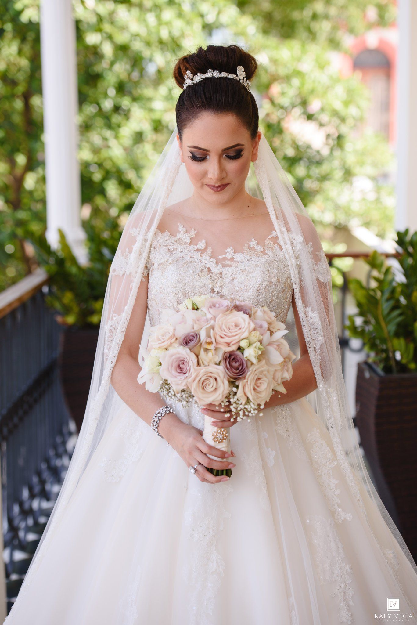 Bride in lace ballgown wedding dress holding blush rose bouquet, wearing tiara and veil, standing outdoors.