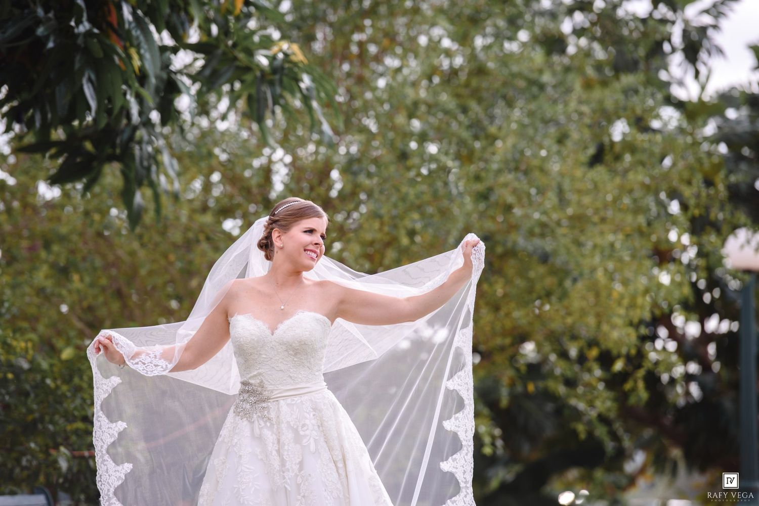 Bride in strapless lace gown holding flowing veil outdoors on wedding day.