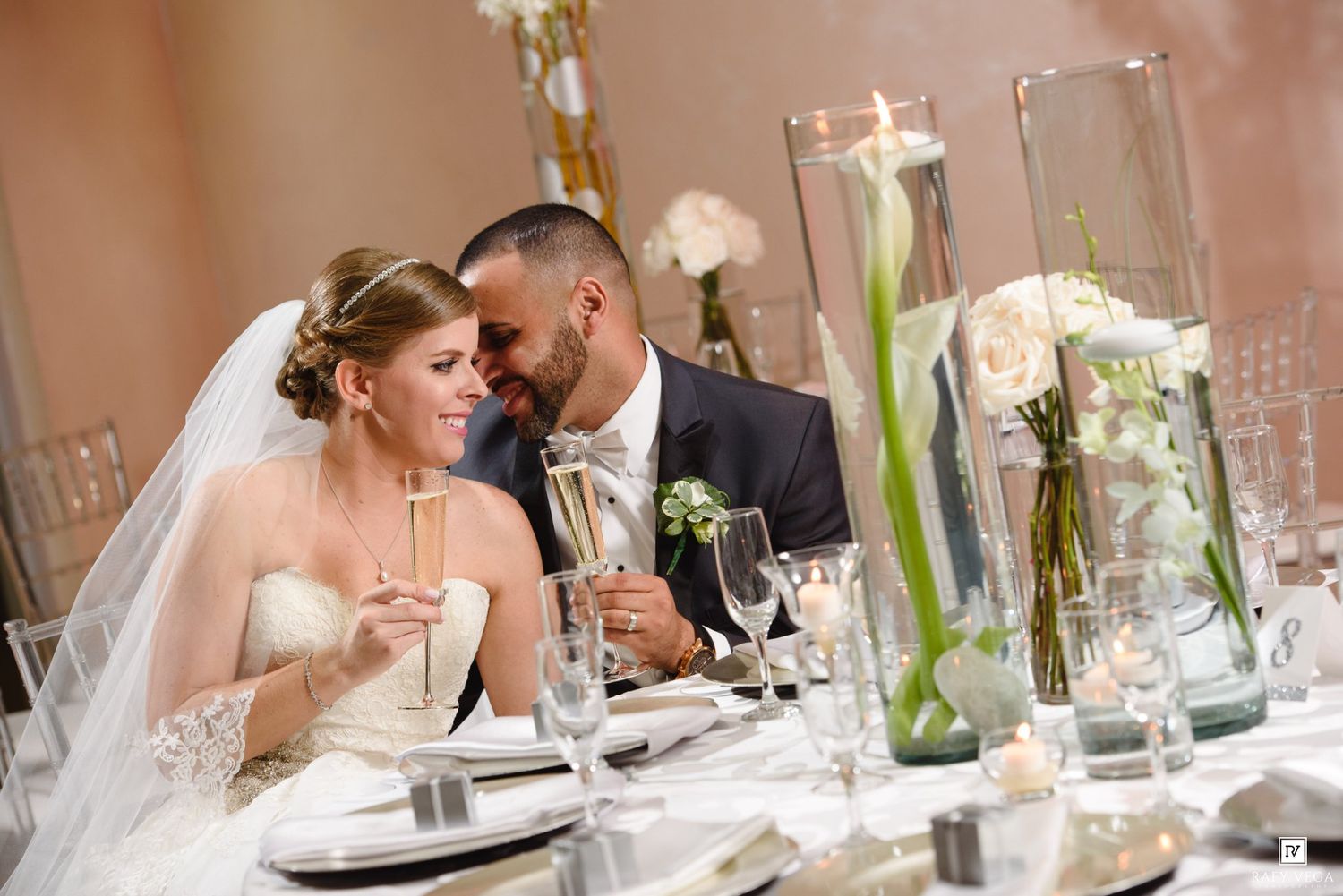 Bride and groom share a romantic moment at their wedding reception table adorned with elegant floral centerpieces and champagne glasses.