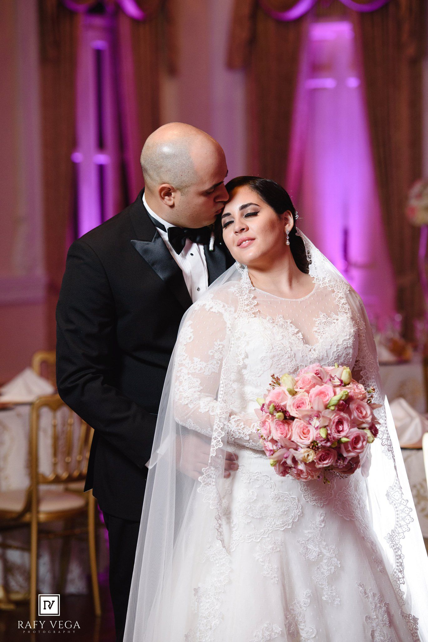 Bride in lace gown holding pink bouquet receives kiss from groom in tuxedo at elegant wedding reception.