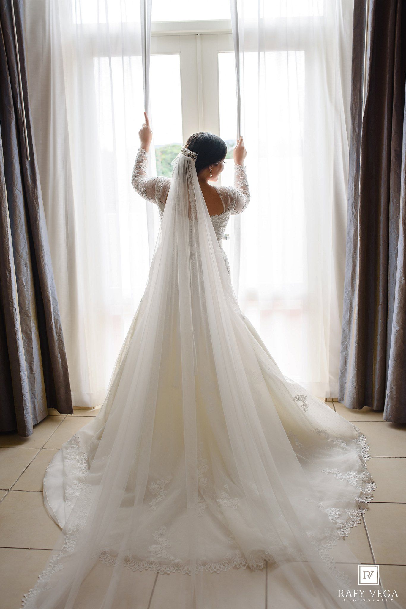 Bride in a white lace wedding gown with long veil stands before a bright window, viewed from behind.