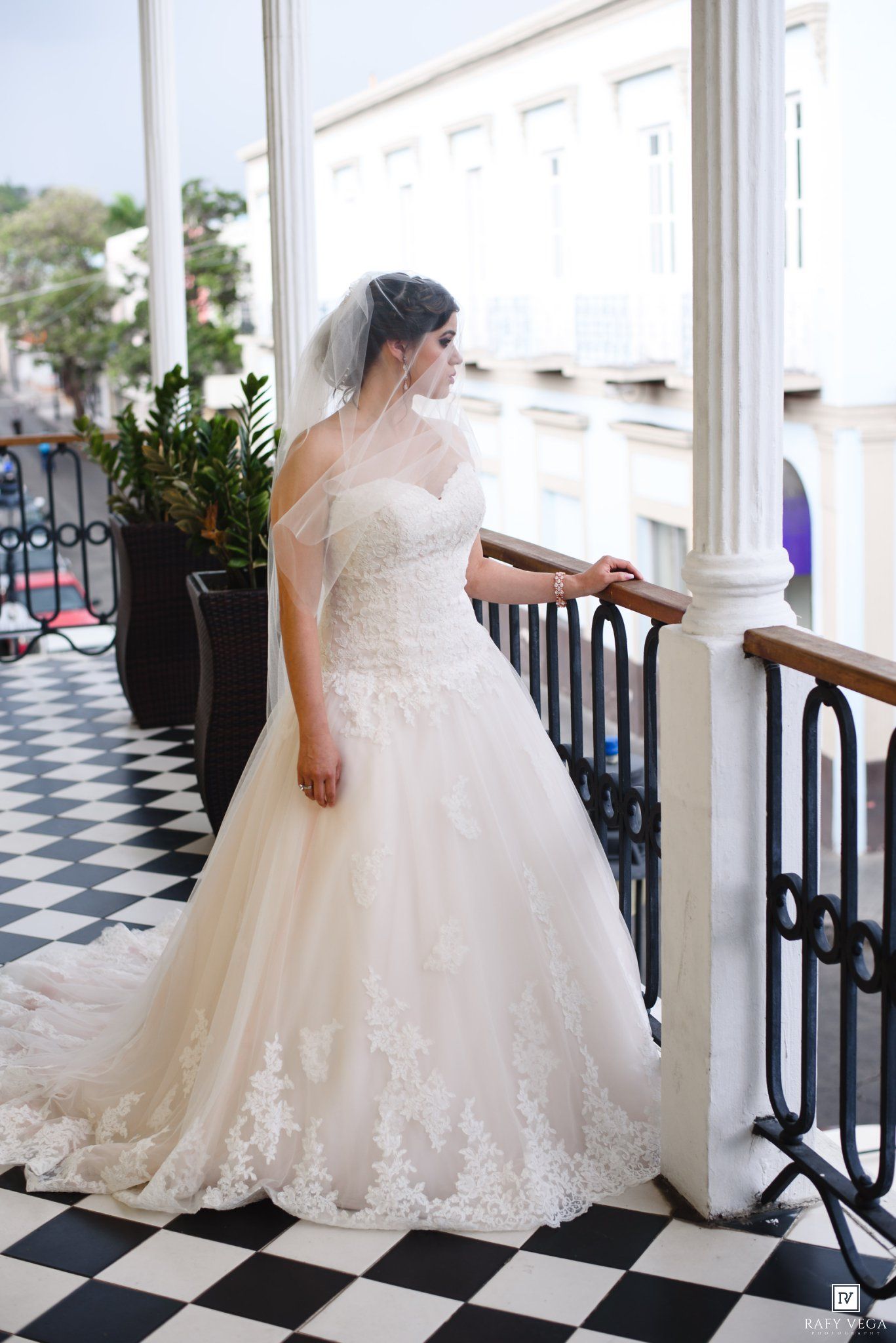 Bride in a white lace ball gown and veil stands on a checkered floor balcony, gazing down elegantly.