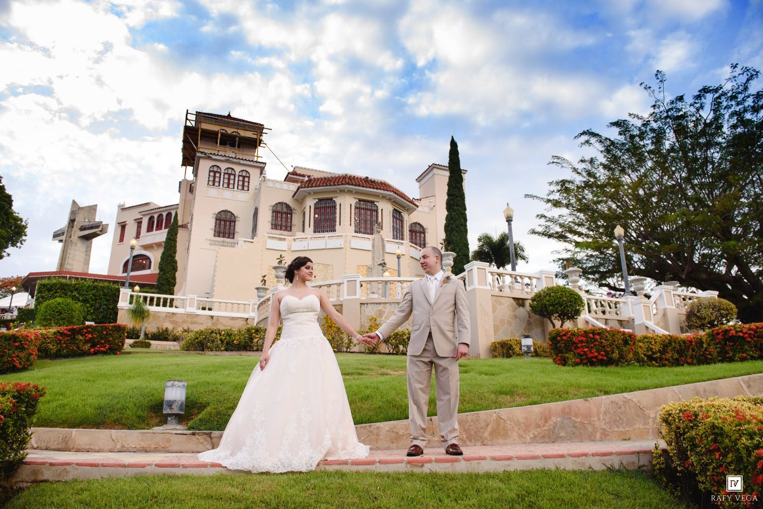 Bride and groom hold hands in front of an elegant Spanish-style mansion surrounded by manicured gardens.