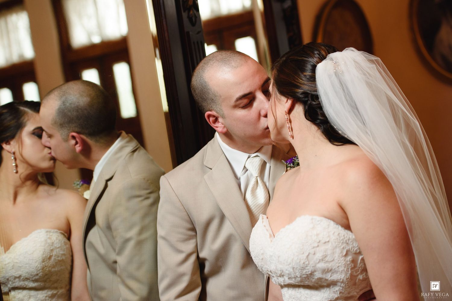 Newlywed couple shares first kiss as bride and groom reflected in mirror at romantic wedding reception.