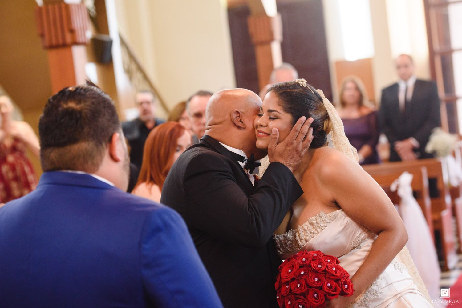 Groom kisses bride holding red bouquet at wedding ceremony altar in a church.