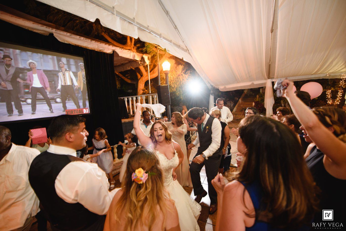 Bride in white gown dancing joyfully with guests at an outdoor wedding reception under a white tent.