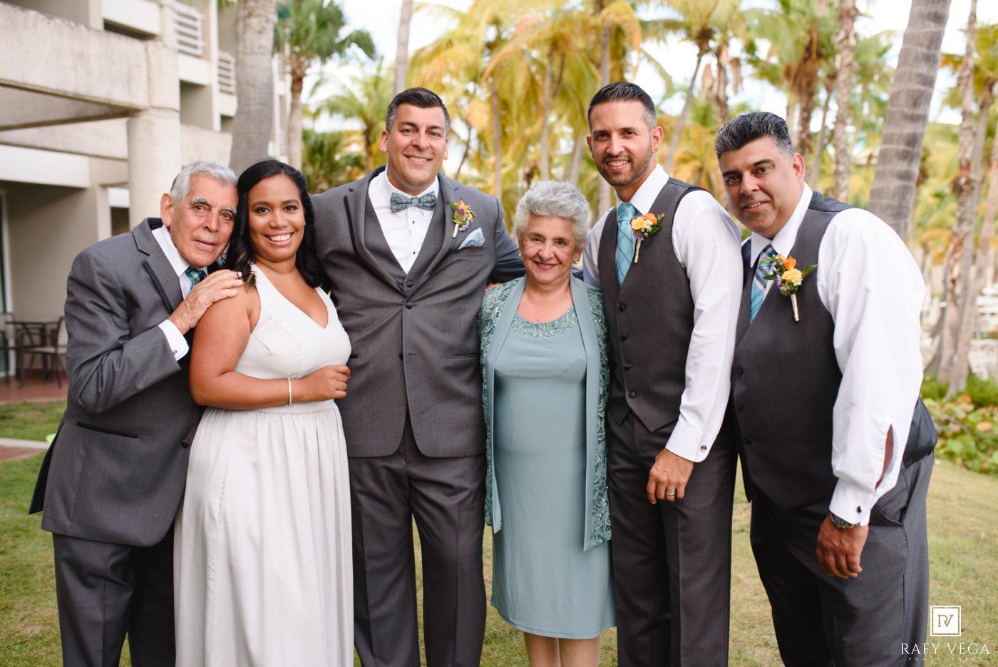 Wedding group photo of six people posing outdoors, including bride in white gown and groom in gray suit, with palm trees.