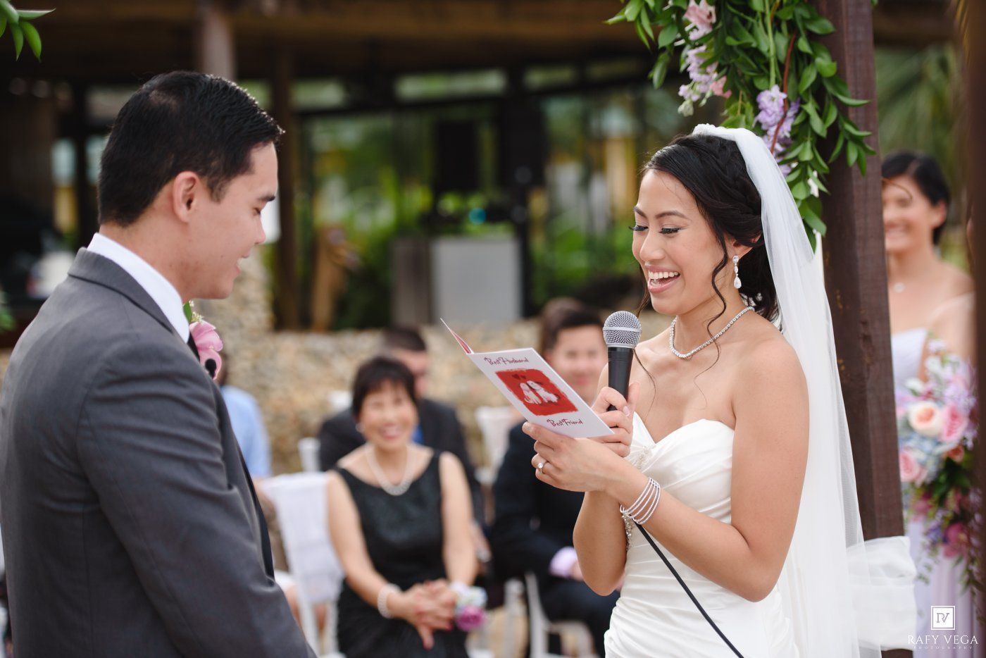 Bride reads wedding vows from a red book into a microphone while smiling at groom during outdoor ceremony.
