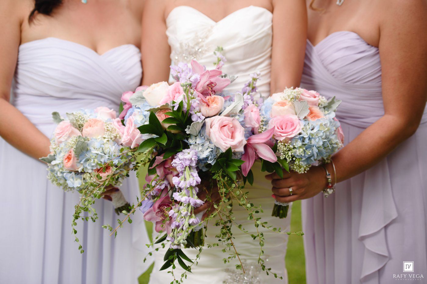 Bride and bridesmaids in lavender dresses holding colorful wedding bouquets with pink roses and blue hydrangeas.