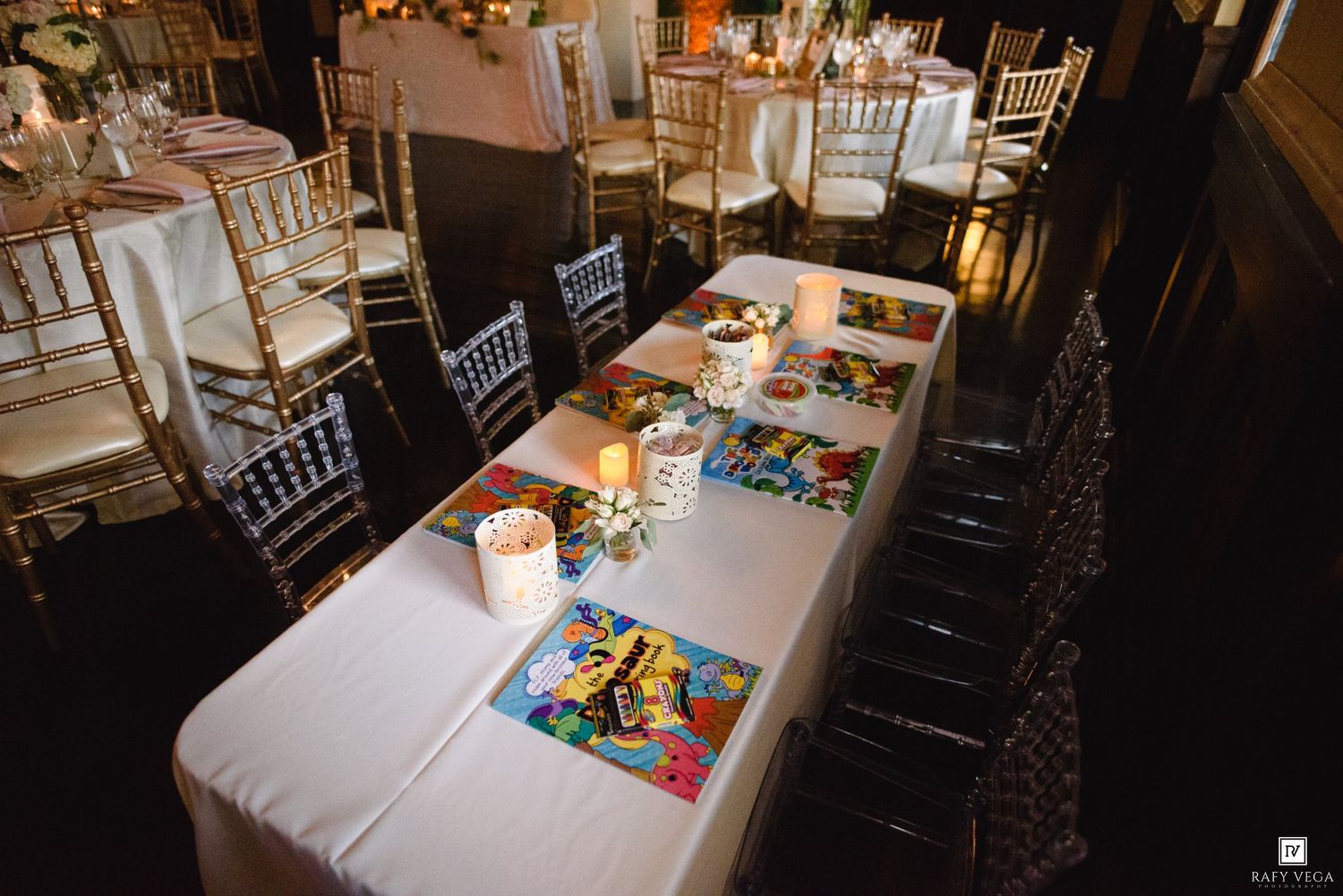 Elegant wedding reception venue with white tablecloths, colorful placemats, gold chiavari chairs, and candlelit round tables in background.