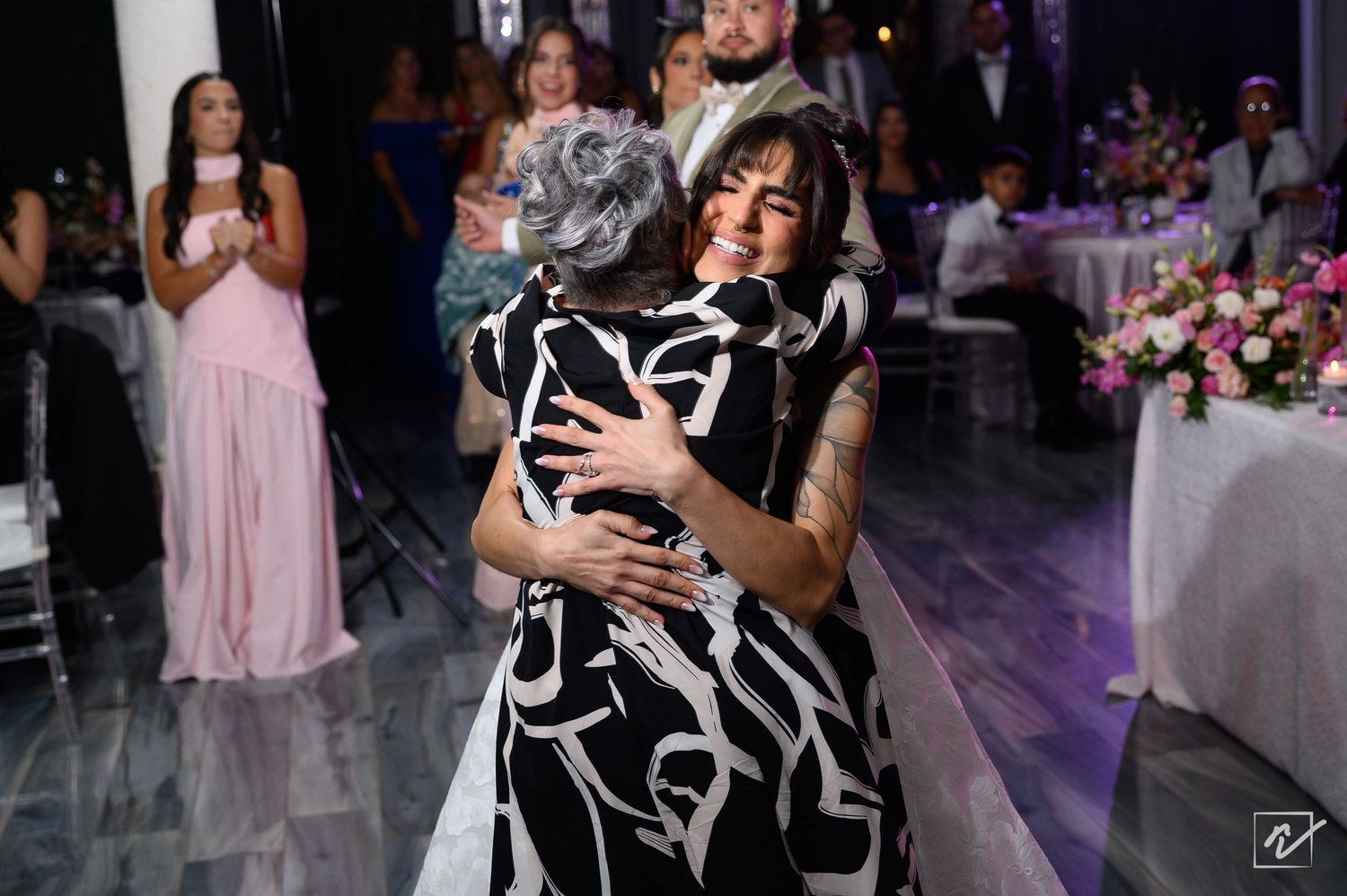 Two women in a tight, emotional embrace on the dance floor at a wedding reception, with guests and floral centerpieces in the background.