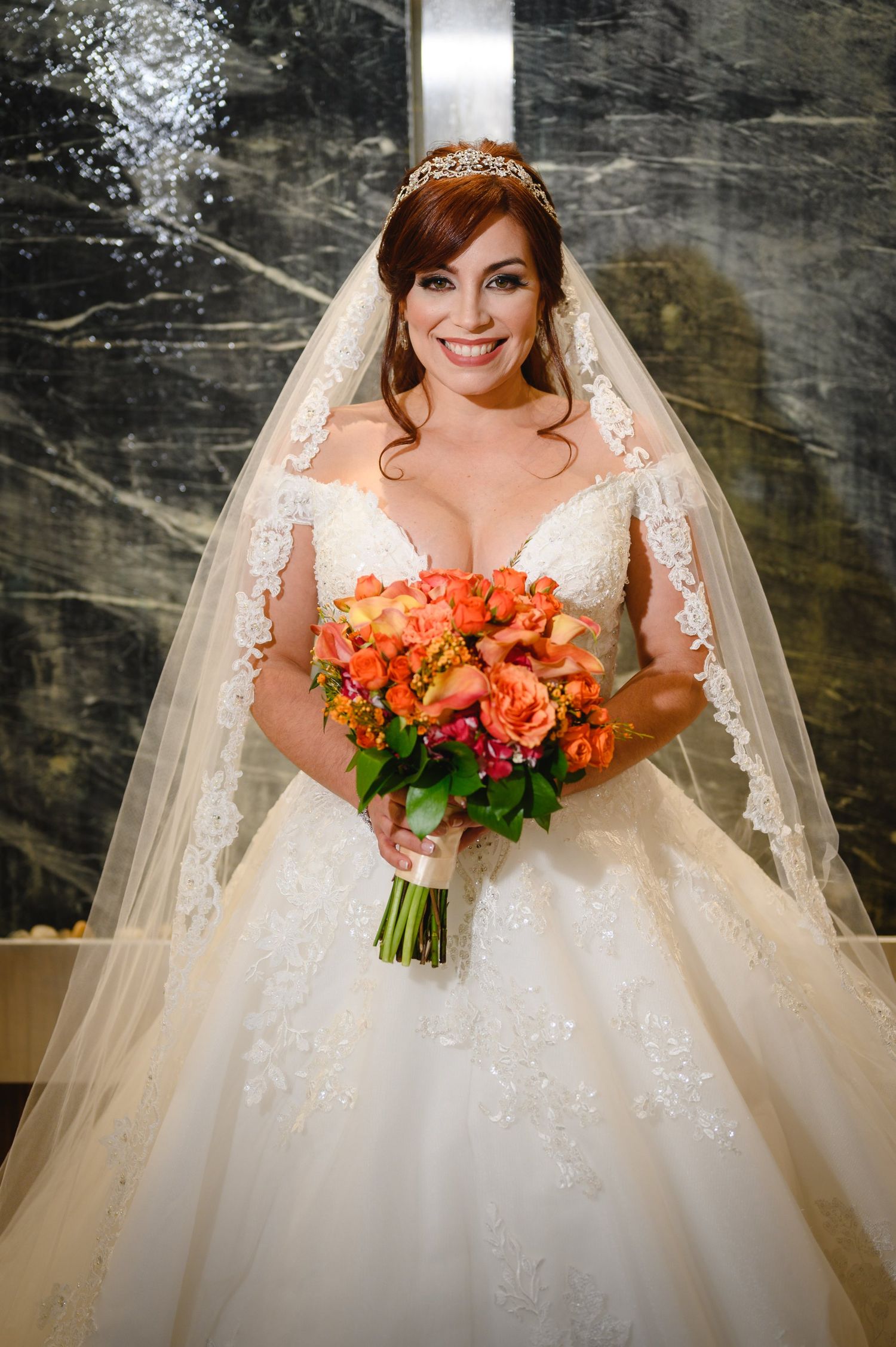 Smiling bride in lace wedding gown and tiara holding vibrant orange floral bouquet in marble setting.